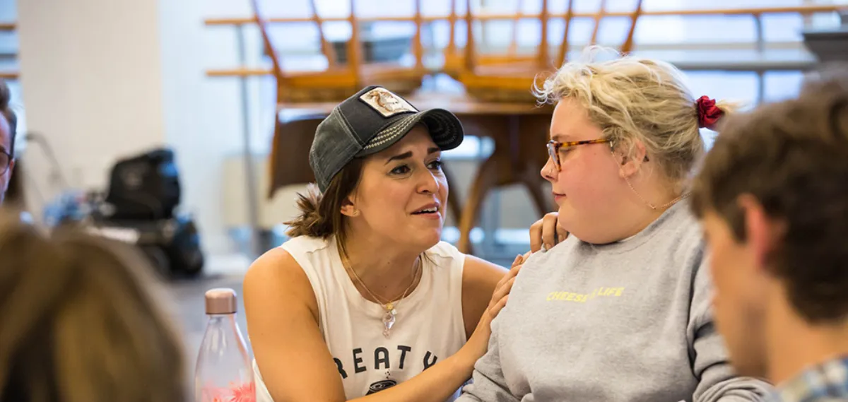A woman in a cap speaks emotionally to another woman wearing glasses and a sweatshirt, placing her hand on the other's shoulder in a supportive gesture.