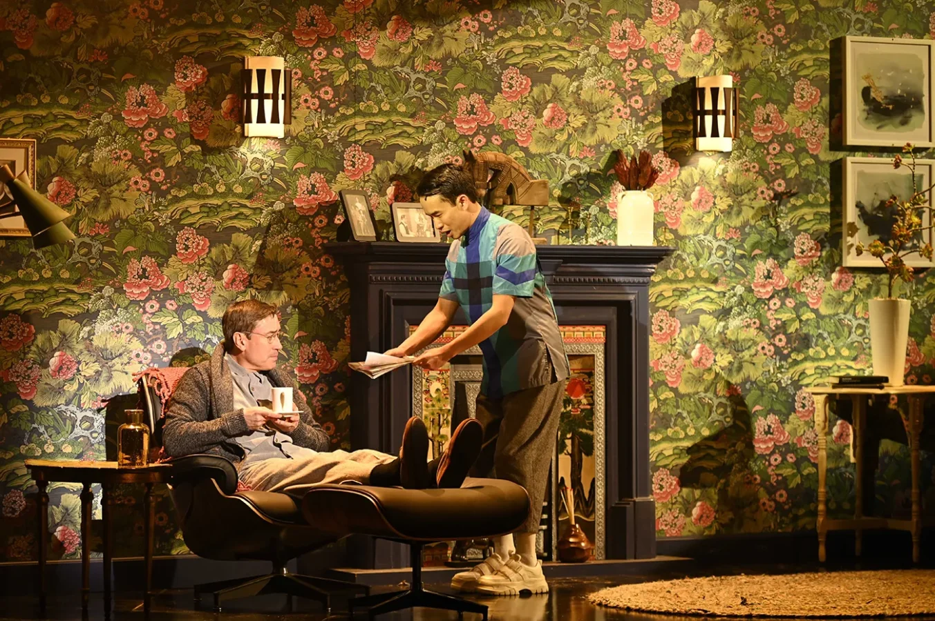 Two men in a room with floral wallpaper; one sits in a lounge chair holding a mug while the other stands, handing him a piece of paper near a fireplace.