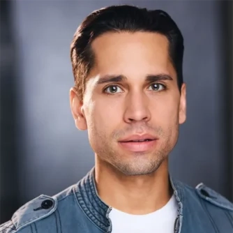 A man with short dark hair, wearing a light blue jacket over a white shirt, looks directly at the camera with a neutral expression.