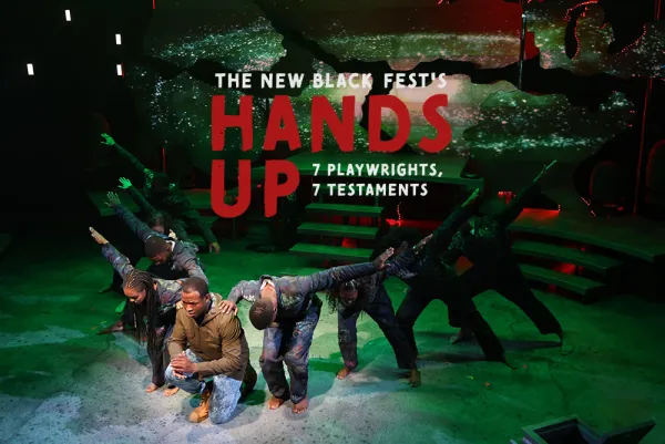 A group of performers on stage, some kneeling and others standing with arms raised, under a sign reading "The New Black Fest's Hands Up: 7 Playwrights, 7 Testaments.