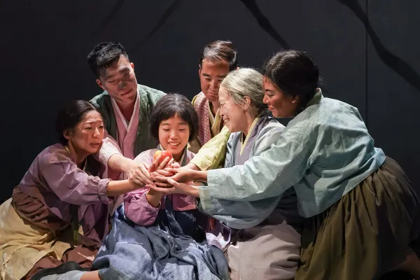 Six people in traditional Korean attire sit closely together, reaching toward the hands of a smiling young woman at the center, sharing an intimate, supportive moment.