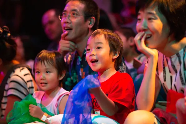 Three children and two adults watch an event, smiling and looking excited. The children hold colorful translucent fabric. The group is seated in a lively, brightly lit setting.