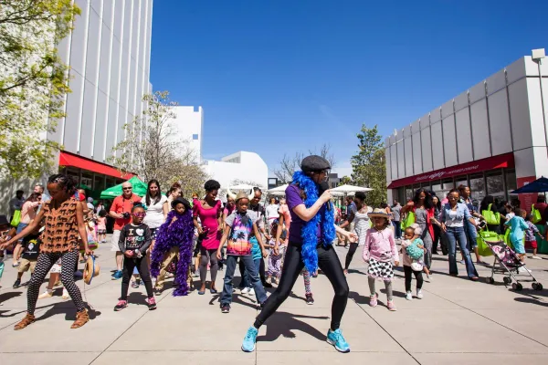 A group of people, including adults and children, participate in a coordinated outdoor dance event on a sunny day in an urban area.