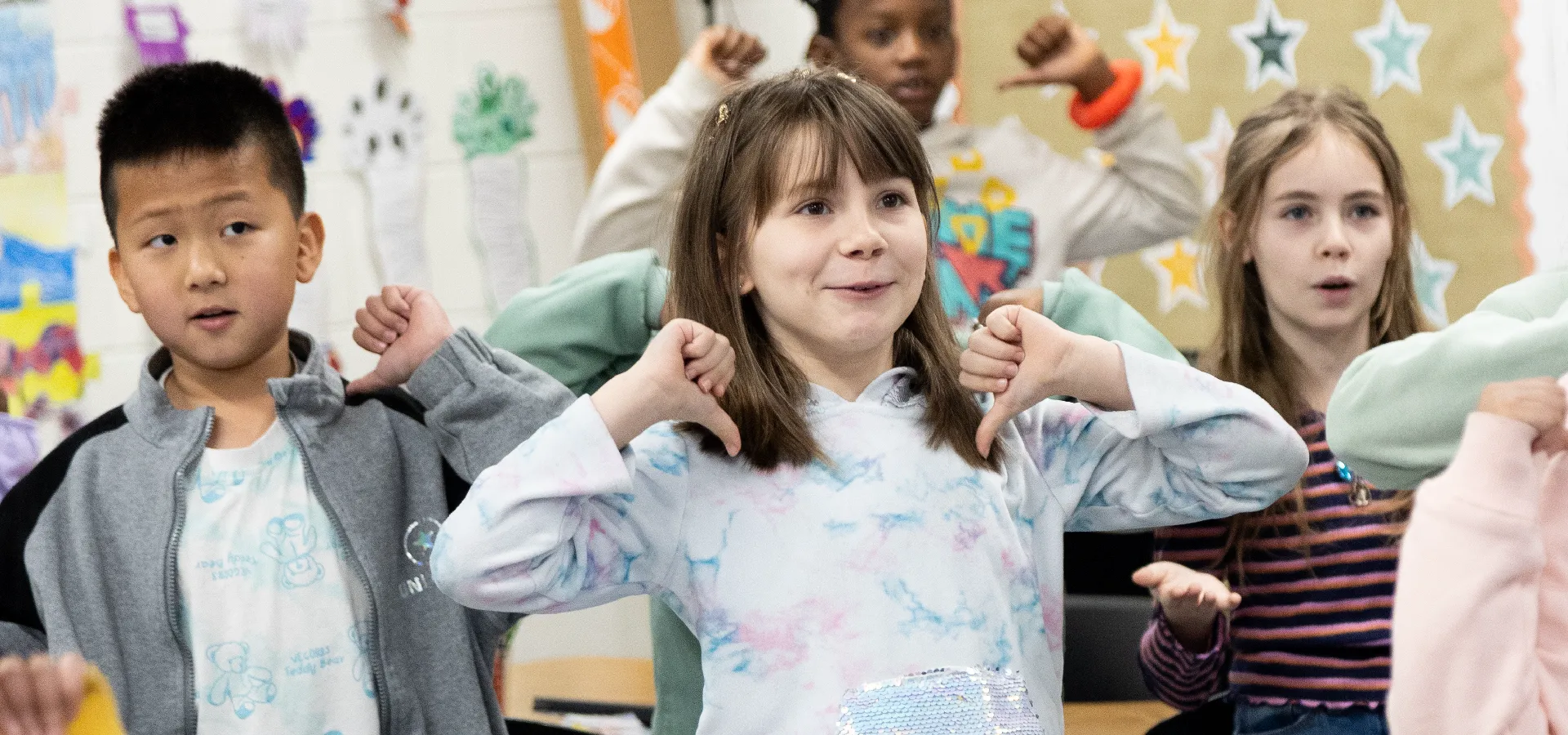 A group of elementary school children stand in a classroom, raising their arms and smiling as they participate in an activity together.