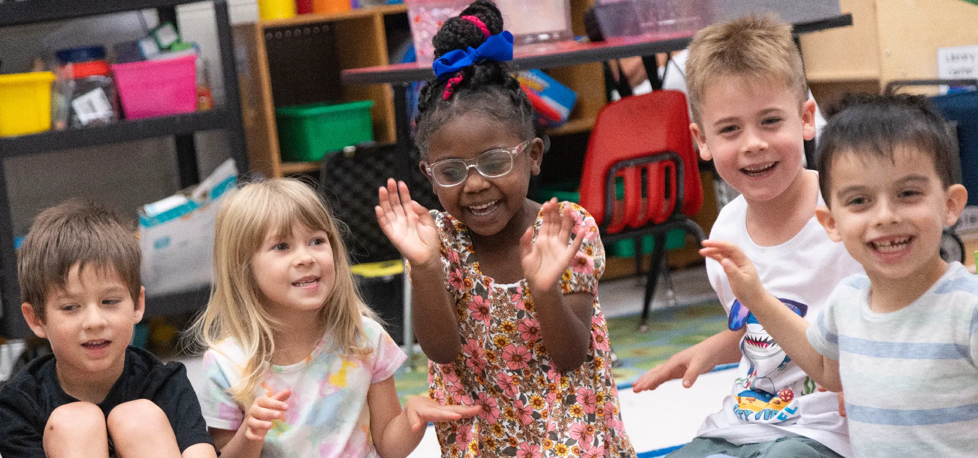 Five young children sit on the floor indoors, smiling and clapping. Shelves with colorful bins are visible in the background.