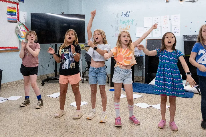 Five children stand in a classroom singing or performing with expressive gestures; papers are scattered on the floor and a TV is in the background.