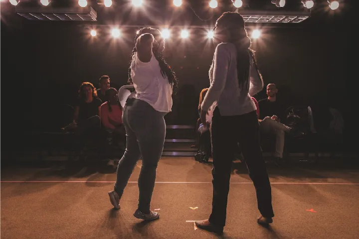 Two people stand and face each other on a stage with bright lights overhead, while an audience watches from the background.