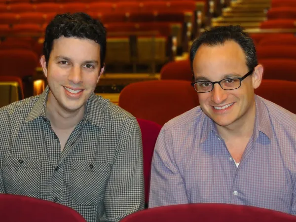 Two men in checkered shirts sit in red theater seats, smiling at the camera. The background shows more empty rows of seats.