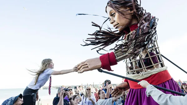 A large puppet interacts with a young girl reaching out her hand, while a crowd of people watches outdoors near the water.