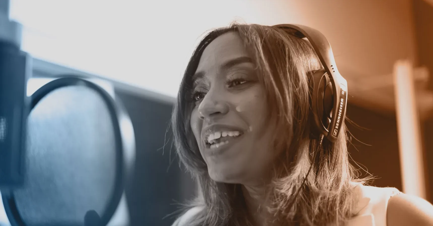A woman wearing headphones is smiling and speaking or singing into a microphone in a recording studio.