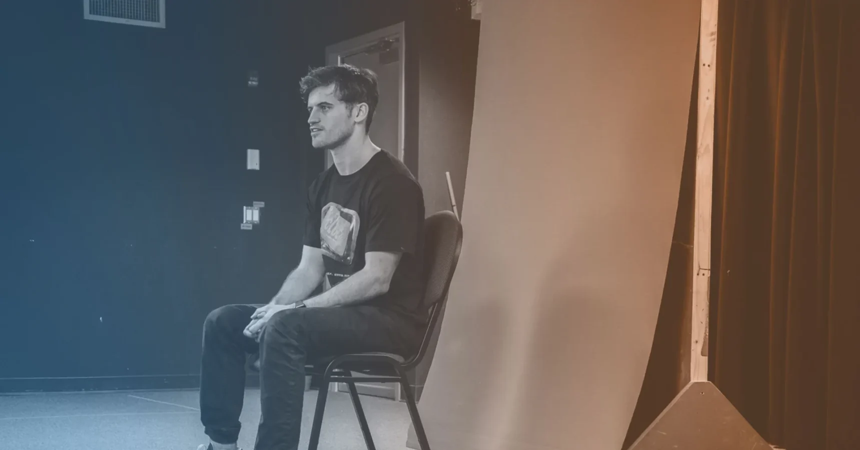A man sits on a chair in a photography studio, facing slightly left. The background includes a gradient backdrop and studio equipment.