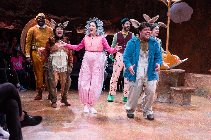 A group of actors in colorful animal costumes perform on stage in front of an audience.