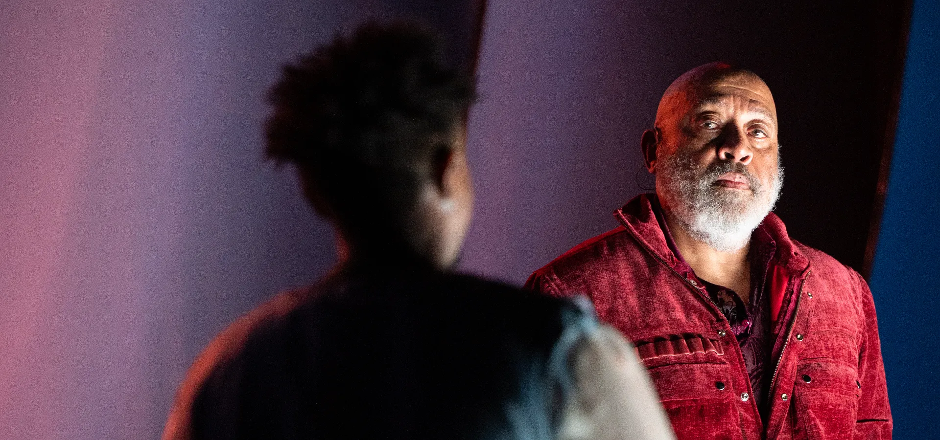 A man in a red jacket with a gray beard looks at another person whose back is facing the camera, against a softly lit background.