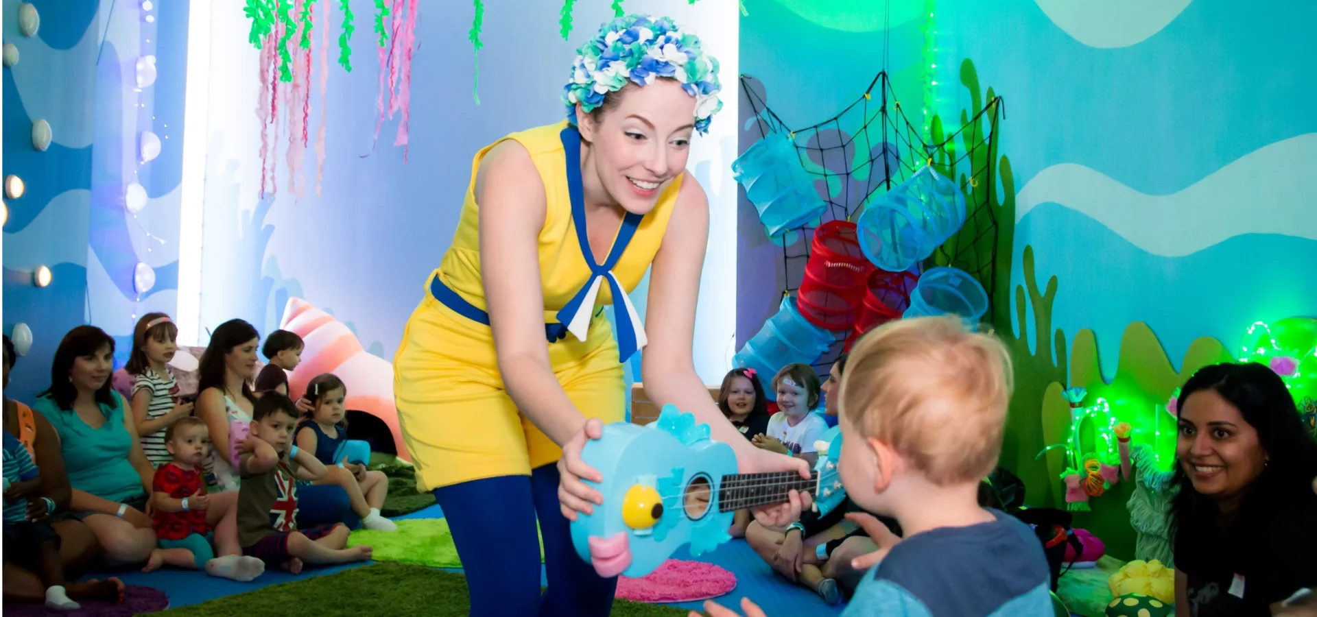 A performer in a yellow dress and blue leggings hands a blue fish-shaped guitar to a young child in a colorful room with an audience of children and adults.