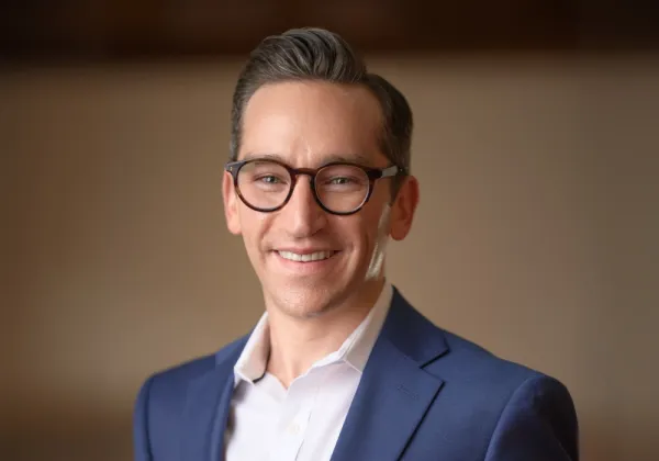 A man with short brown hair and glasses, wearing a blue suit jacket and white shirt, smiles at the camera against a plain background.