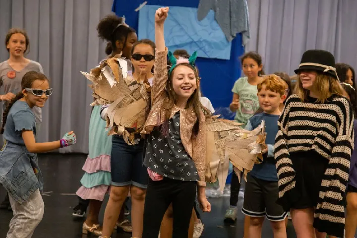 A group of children stand together indoors; one girl in the center raises her fist and wears a costume with gold wings. Others around her wear casual clothes and some have sunglasses or hats.