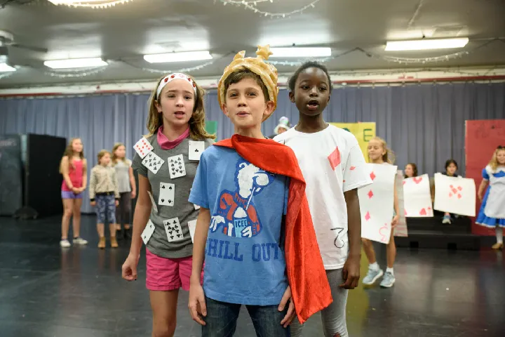 Three children in homemade playing card costumes stand at the front of a stage, with other kids and simple props visible in the background, suggesting a school play or rehearsal.