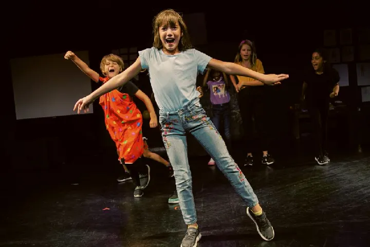 A group of children energetically dance and pose on a dimly lit stage, with one girl in a light shirt and jeans at the front.