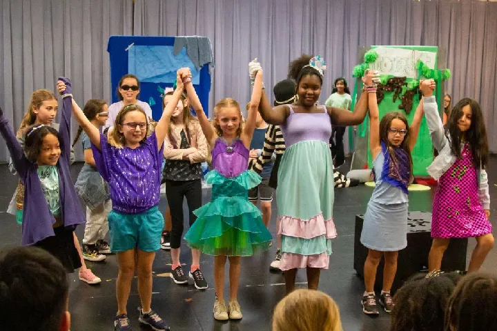 A group of children stand on stage holding hands and raising their arms, with some dressed in colorful costumes, while others watch in the background.