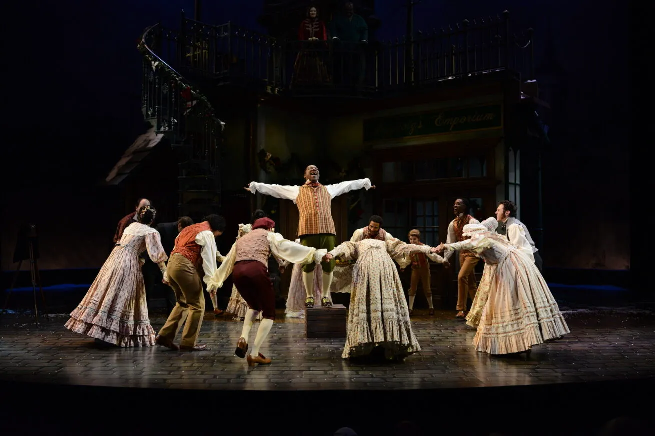 A group of performers in period costumes dance in a circle on a theater stage, with one person standing on a platform at the center.