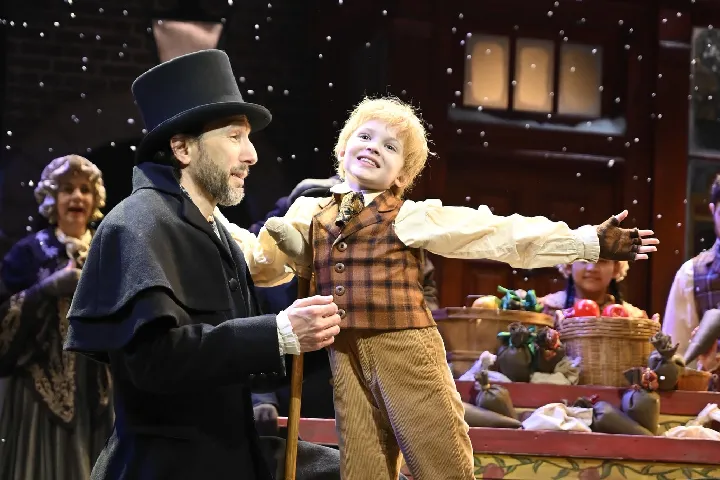 A man in a black coat and top hat kneels beside a smiling child in period costume with arms outstretched, on a festive stage with baskets of produce and artificial snow falling.
