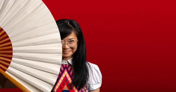 A woman with long black hair and glasses stands against a red background, partially hiding her face behind a large white fan.