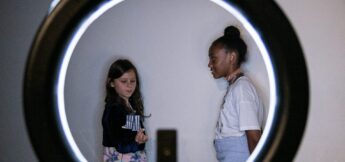 Two girls stand against a white wall, one holding a prop, viewed through the circular frame of a ring light.