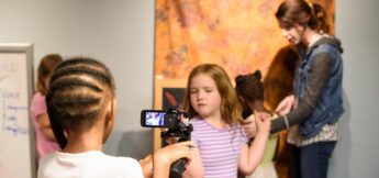 A child holds a video camera, filming other children and an adult during a Camera Acting class in a classroom.