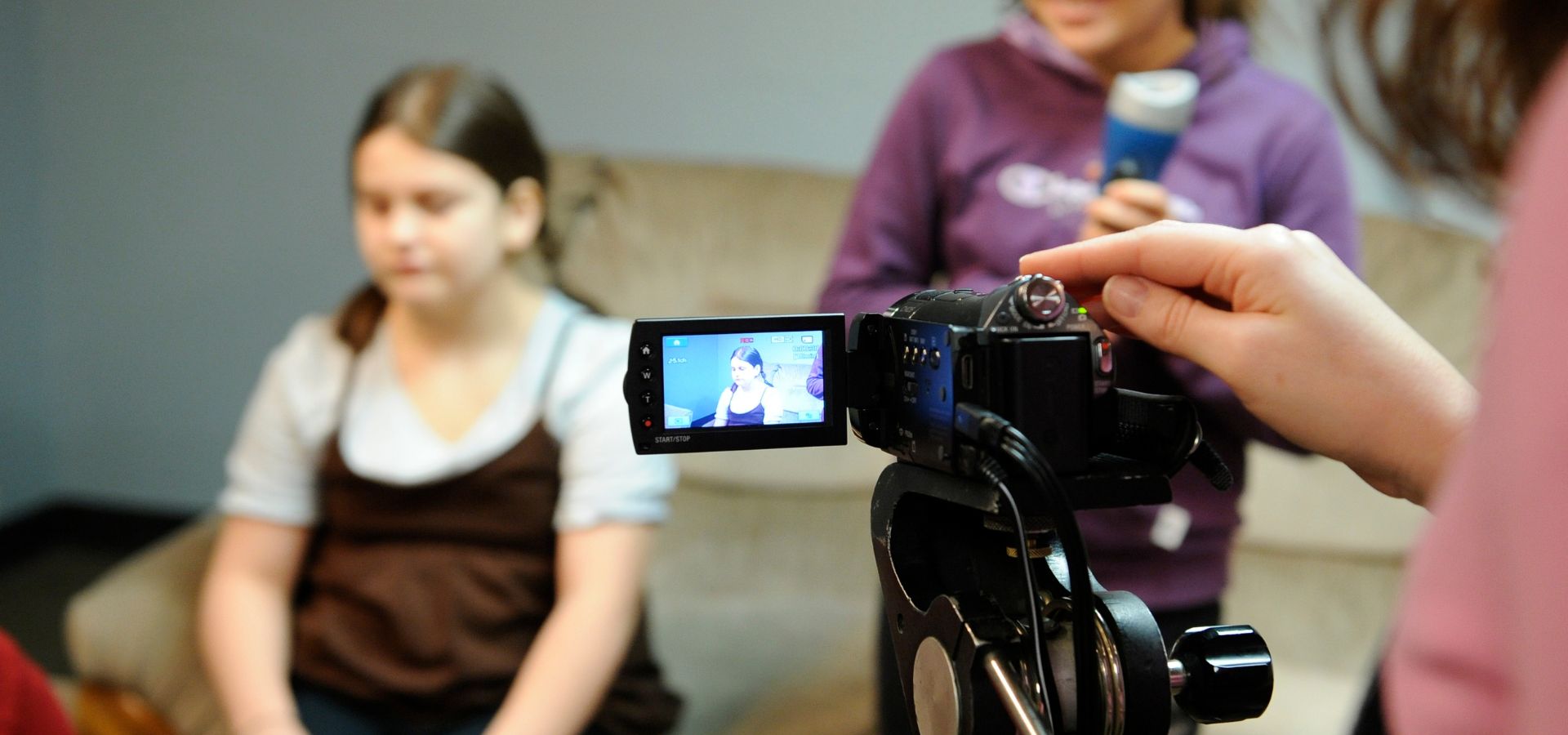 A person records a young girl sitting on a couch while another person stands nearby holding an object, possibly a microphone.