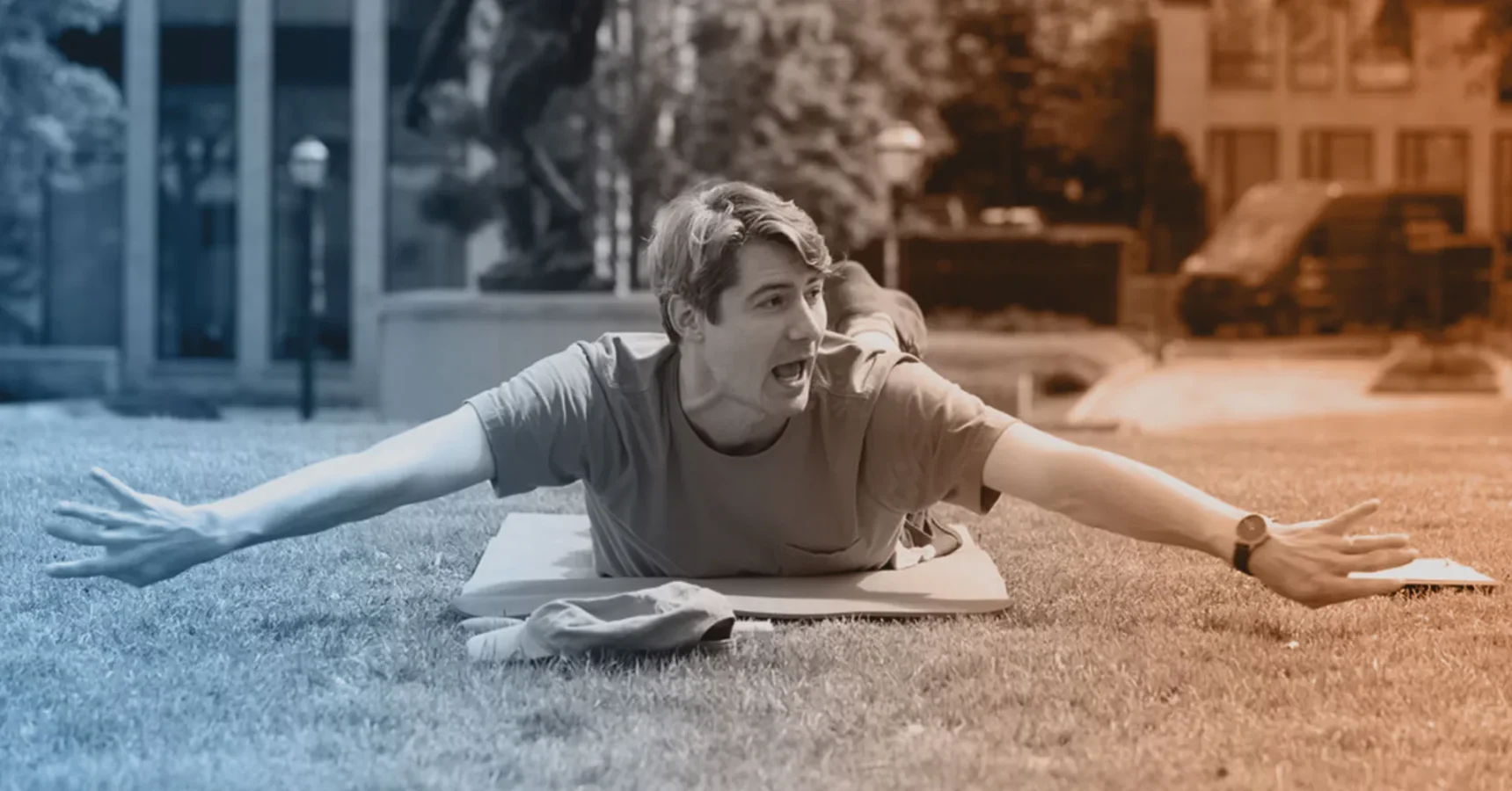 A man lies on his stomach on a mat outdoors with arms outstretched and mouth open, appearing to shout or express excitement.