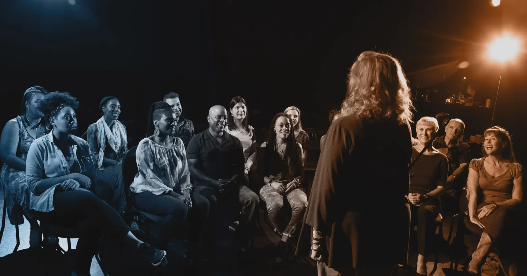 A person stands and speaks on stage to a seated audience, who are watching and reacting, in a dimly lit setting.
