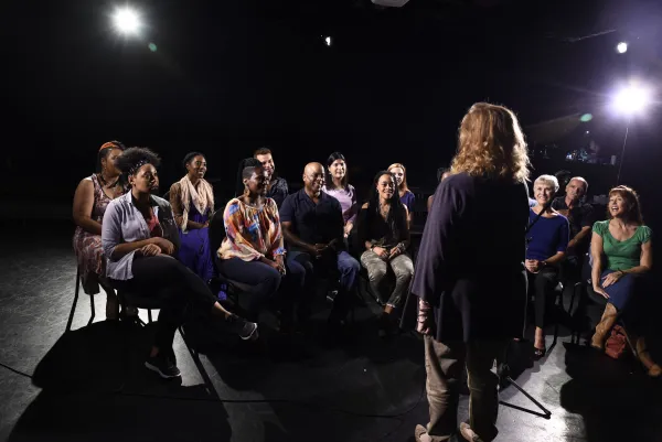 A person stands and speaks to a seated group of adults in a dark room with bright stage lights overhead.