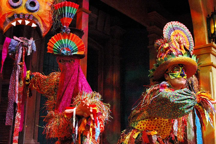 Two performers in colorful, elaborate costumes and masks pose on stage, surrounded by vibrant props and theatrical lighting.