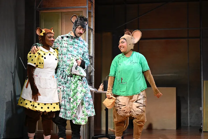 Three actors in mouse costumes perform on stage, wearing playful outfits and standing in front of a set with industrial-style scaffolding and props.