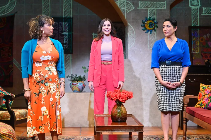Three women stand on a colorful stage set; one wears an orange floral dress, one a pink suit, and one a blue top with a checkered skirt. A vase with red flowers is on a table in front.