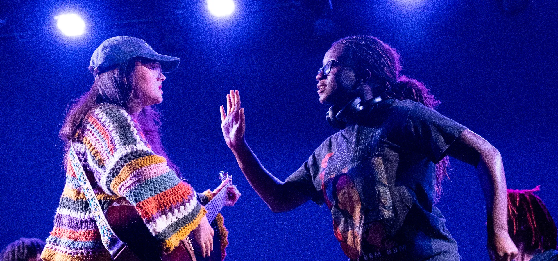 Two people face each other under blue stage lights; one holds a guitar and wears a cap, while the other raises a hand, wearing headphones around their neck.