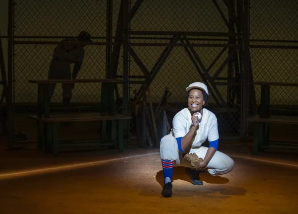 A baseball player in a white uniform crouches and smiles on the field under a spotlight, with another player in the background near a chain-link fence and benches.