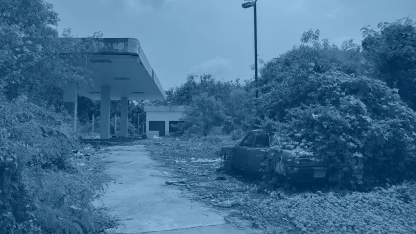 An abandoned gas station with overgrown vegetation and a car partially covered by bushes.