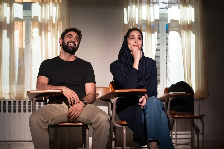 A man and a woman sit next to each other at desks in a classroom with light streaming through curtained windows behind them.