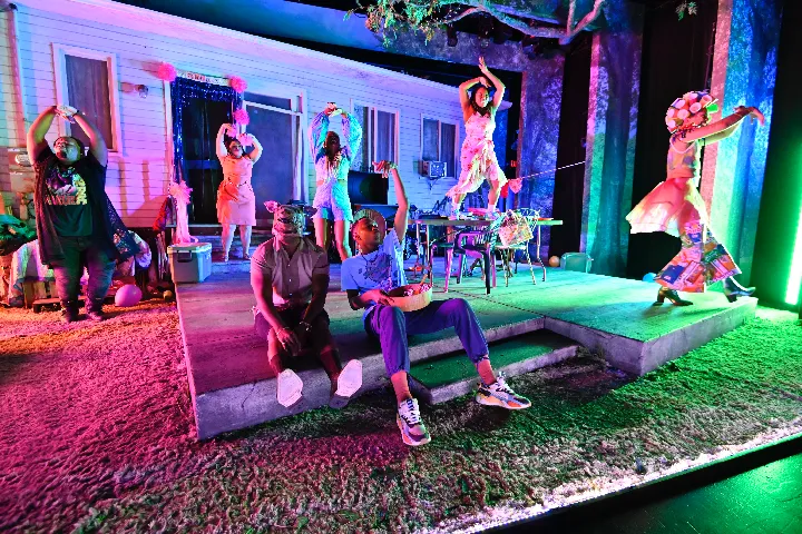 Six people perform on a colorful stage set resembling a house and yard, with two seated in front and four standing or dancing in vibrant costumes under theatrical lighting.