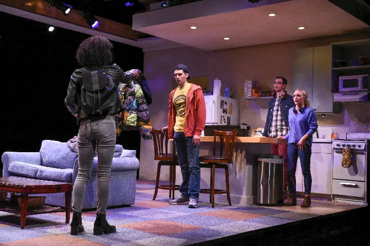 Four people are onstage in a modern living room and kitchen set; one person stands with their back to the camera, facing the other three who watch attentively.
