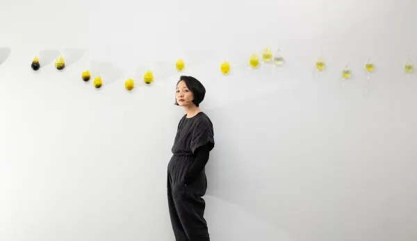 A woman in dark clothing stands against a white wall with a row of glass vessels containing yellow liquid arranged diagonally above her.
