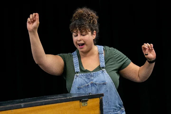 A person wearing a green shirt and denim overalls gestures with both arms raised and mouth open, standing behind a wooden surface against a black background.