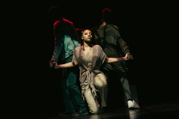 Three performers are on a dimly lit stage; the central figure kneels with arms outstretched, while two others stand with their backs turned.