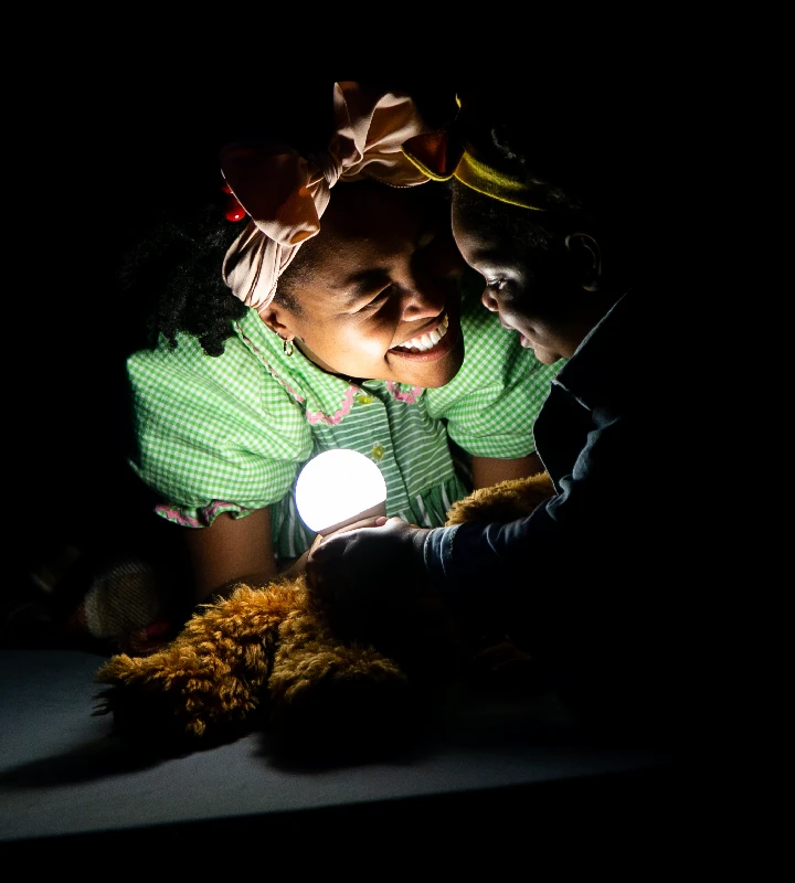 An adult and a child smile at each other while holding a glowing round light in a dark room, with a stuffed toy visible in the foreground.