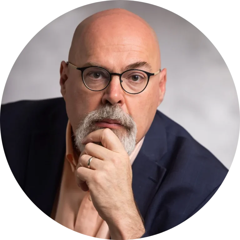 Bald man with glasses and a goatee wearing a dark suit jacket and peach shirt, resting his chin on his hand, looking directly at the camera.