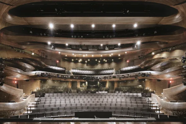 A modern, multi-tiered theater auditorium with wooden accents, empty gray seats, and bright stage lights facing the camera.
