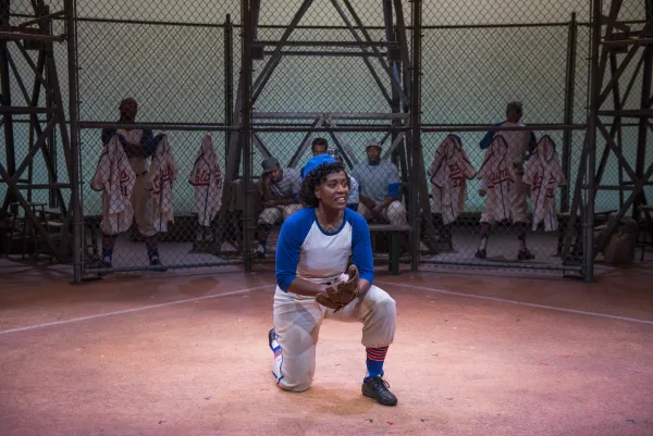 A baseball player in a blue and white uniform kneels on the field holding a glove, with teammates sitting behind a chain-link fence in the background.
