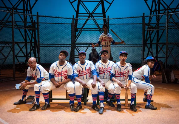 Seven baseball players in matching blue and white uniforms sit and stand in a dugout area with a chain-link fence and metal structure in the background.