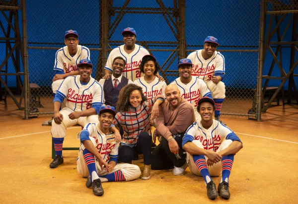 A group of actors in vintage baseball uniforms and casual attire pose together on a baseball field set in front of a chain-link fence.
